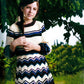 A young girl stands outdoors under a tree, wearing a knitted dress with horizontal stripes and zigzag patterns in various colors from Malabrigo Yarn's Malabrigo Book 9 - Niños. She looks at the camera with a gentle expression, her long hair casually draped over one shoulder. The background includes greenery and a grassy path.