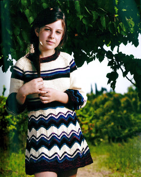 A young girl stands outdoors under a tree, wearing a knitted dress with horizontal stripes and zigzag patterns in various colors from Malabrigo Yarn's Malabrigo Book 9 - Niños. She looks at the camera with a gentle expression, her long hair casually draped over one shoulder. The background includes greenery and a grassy path.