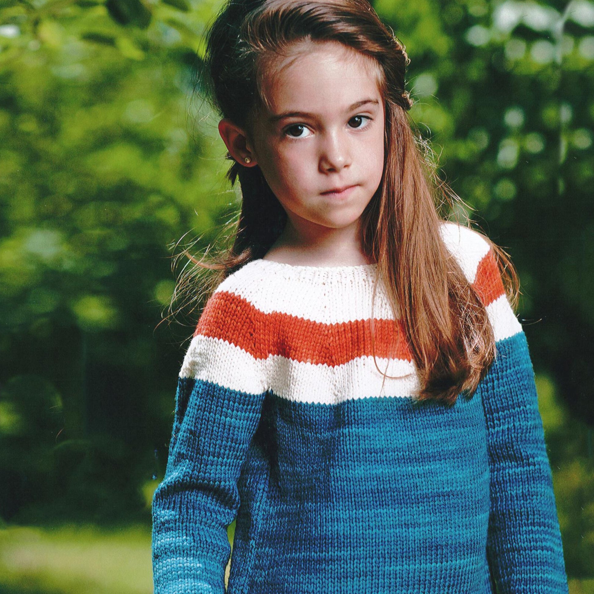 A young girl stands outdoors with a calm expression. She has long brown hair and is wearing a knitted sweater with wide stripes in blue, white, and orange—a design inspired by the unique patterns for kids found in Malabrigo Yarn's Malabrigo Book 9 - Niños. The background is filled with lush green foliage.