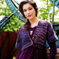 A woman with long dark hair stands outdoors near a sundial, wearing a textured, multicolored knit shrug from Malabrigo Book 11 - Aniversario by Malabrigo Yarn over a purple top. She accessorizes with a necklace and earrings, and greenery is visible in the background.