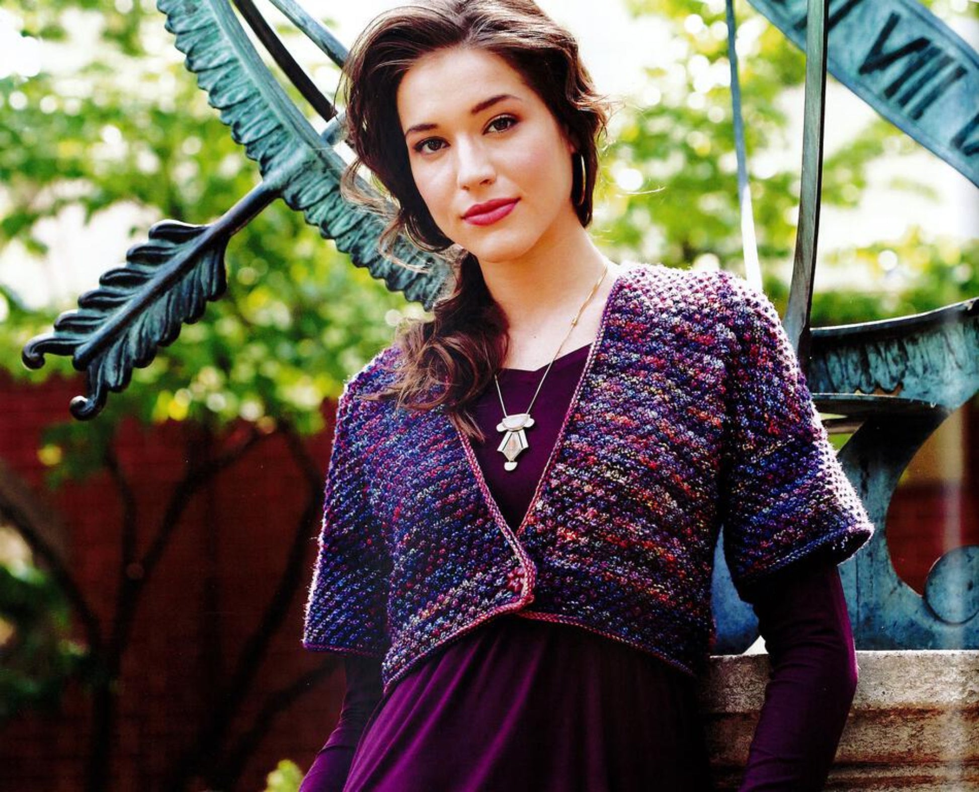 A woman with long dark hair stands outdoors near a sundial, wearing a textured, multicolored knit shrug from Malabrigo Book 11 - Aniversario by Malabrigo Yarn over a purple top. She accessorizes with a necklace and earrings, and greenery is visible in the background.