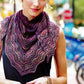 A woman with light-colored hair is wearing a black sleeveless top and a hand-knitted, multicolored lace scarf from Malabrigo Yarn's Malabrigo Book 11 - Aniversario collection around her neck. She is outdoors, with blurred trees and urban elements in the background. The Aniversario shades add vibrancy to her subtle smile as she holds her hands together in front.