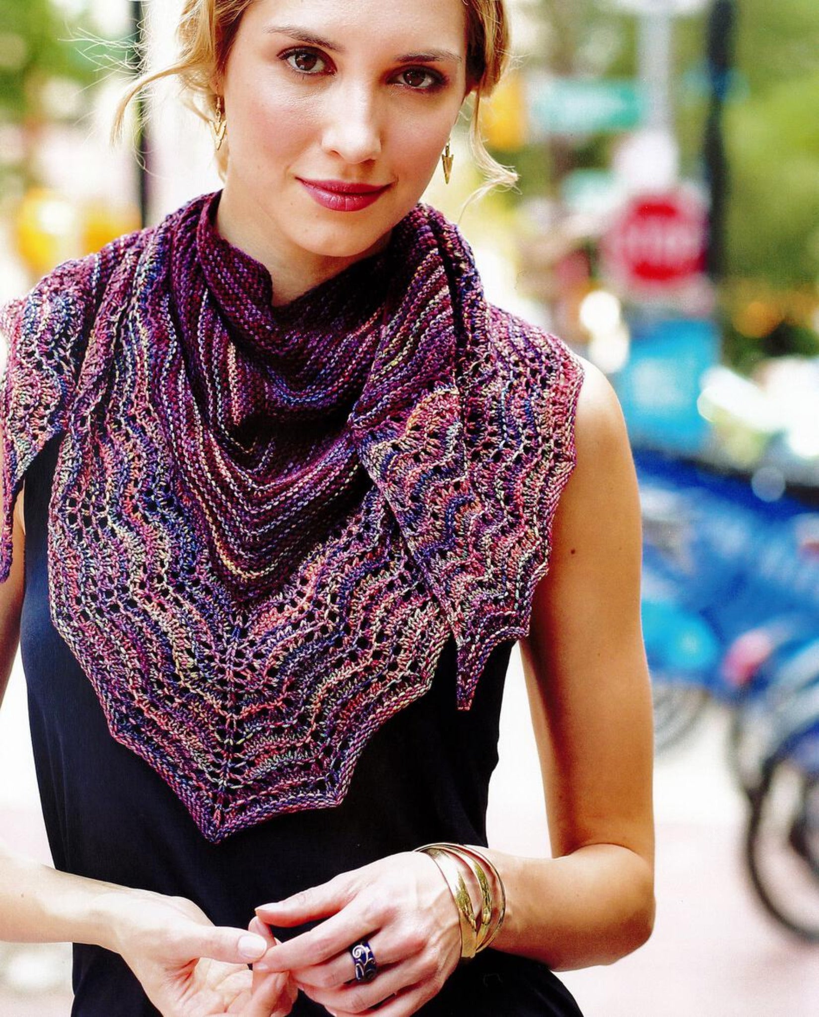 A woman with light-colored hair is wearing a black sleeveless top and a hand-knitted, multicolored lace scarf from Malabrigo Yarn's Malabrigo Book 11 - Aniversario collection around her neck. She is outdoors, with blurred trees and urban elements in the background. The Aniversario shades add vibrancy to her subtle smile as she holds her hands together in front.