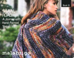 A woman is wearing a multicolored knitted shawl featuring a zigzag pattern, crafted from Malabrigo yarns. The title on the image reads, "Shawl Road: A Journey with Hand-Painted Yarns," which matches the product Malabrigo Book 13 - Shawl Road by Malabrigo Yarn.