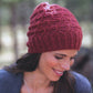 A person with long, dark hair is wearing a red knit beanie from New American Knits by Ingram Content. The beanie features a textured pattern. They are looking down and smiling, dressed in an elegant gray sweater, with a blurred outdoor setting in the background.