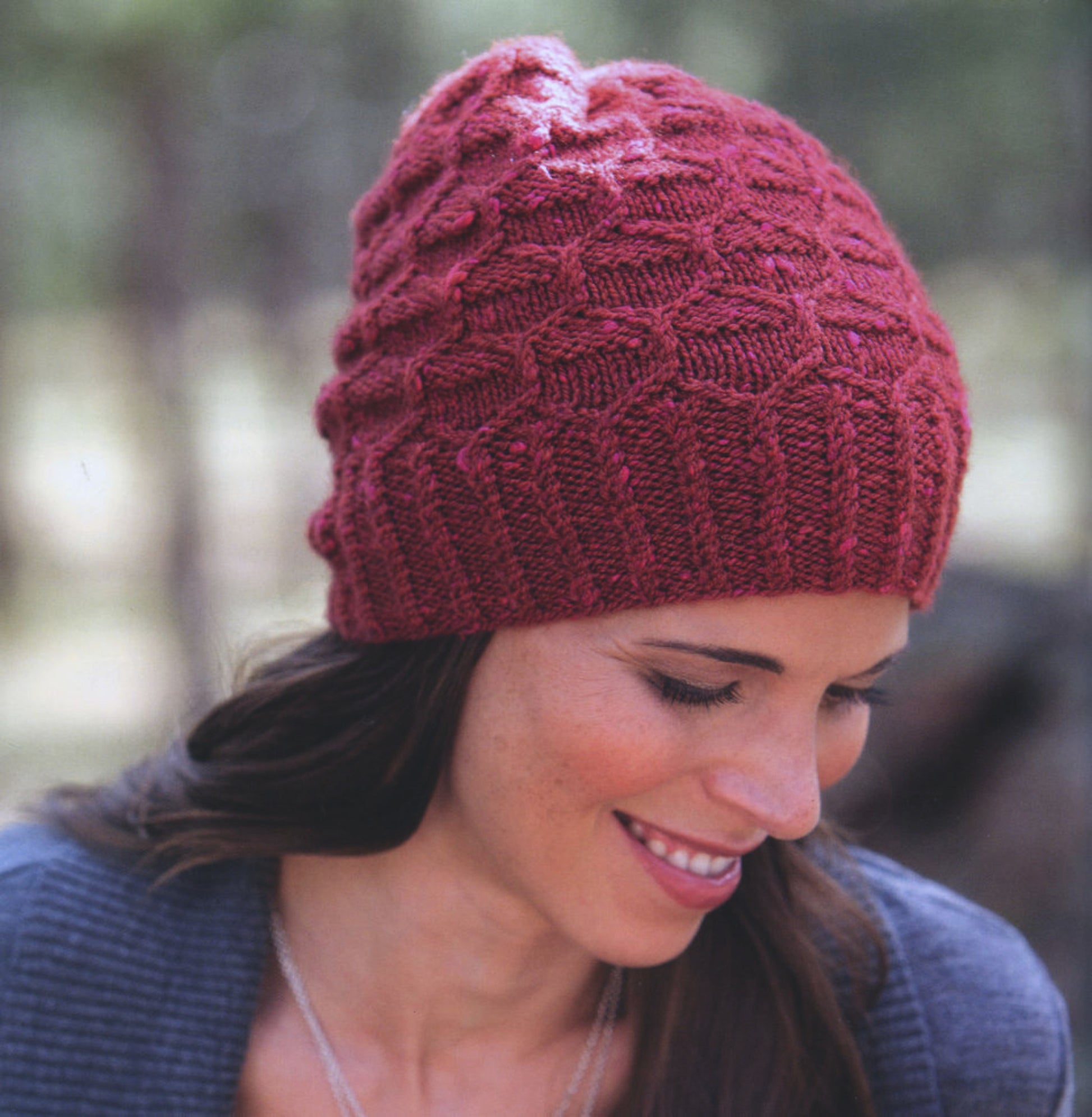 A person with long, dark hair is wearing a red knit beanie from New American Knits by Ingram Content. The beanie features a textured pattern. They are looking down and smiling, dressed in an elegant gray sweater, with a blurred outdoor setting in the background.