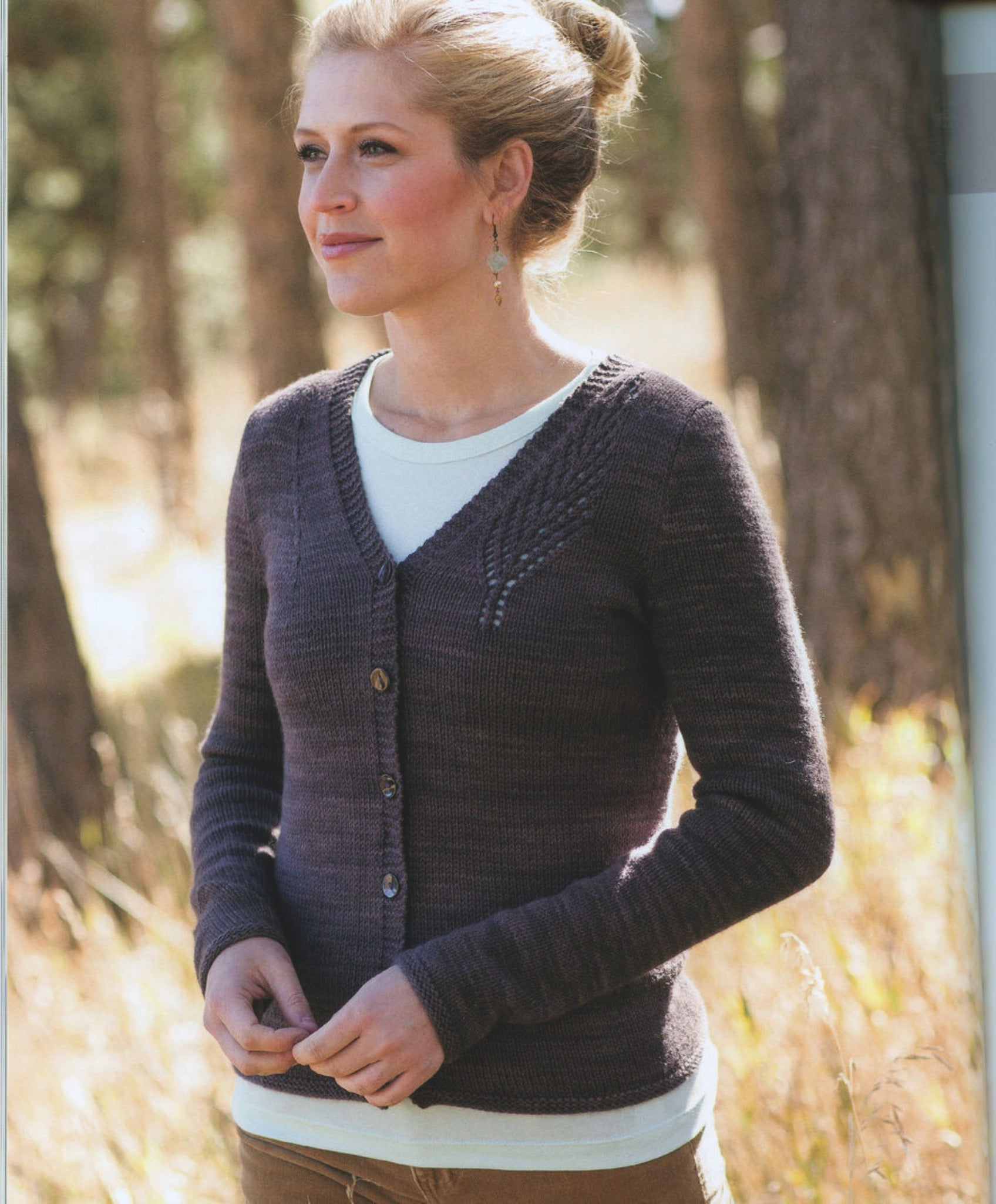 A woman with a light complexion and hair tied in a bun stands outdoors in a wooded area, wearing an elegant sweater with buttons over a white top and earrings, looking slightly to her left. The background features blurred trees and dry grass, capturing the serene setting reminiscent of American artists' favorite landscapes. She is showcasing the "New American Knits" by Ingram Content.