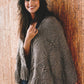 A smiling person stands against a cork wall, draped in the exquisite "New American Knits" poncho from Ingram Content. The poncho boasts intricate decorative patterns and a cozy texture akin to elegant sweaters. They have long, dark hair and are gently holding the edge of the poncho with one hand, looking relaxed and content.