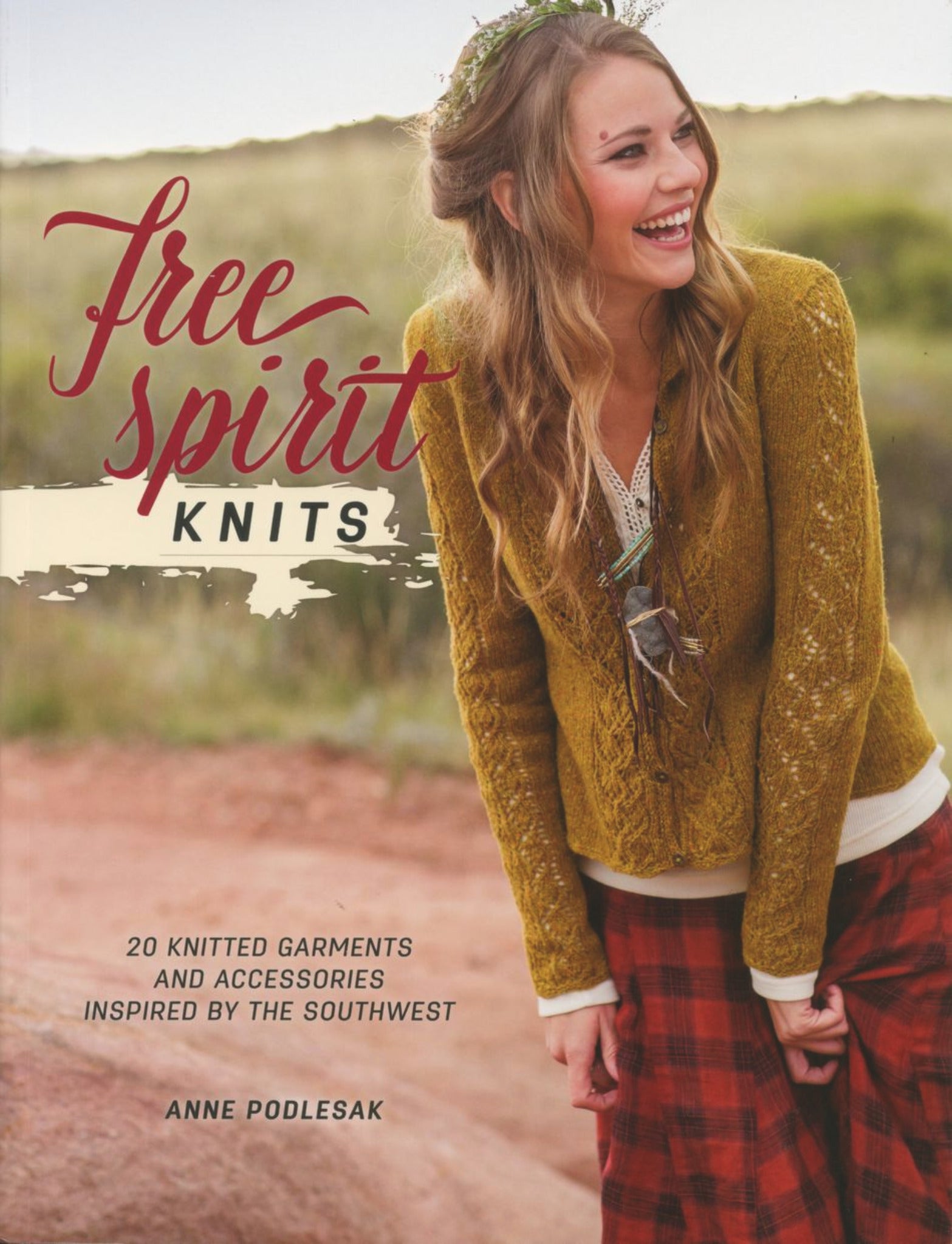 A woman is smiling on the cover of "Free Spirit Knits" by Anne Podlesak, a book published by Ingram Content. She is wearing a knit sweater over a white shirt and red plaid pants. The subtitle reads, "20 knitted garments and accessories inspired by the Southwest," making it perfect for knitters of every skill level.