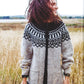 A person with curly hair stands in a field of tall grass, smiling and looking to the side. They are wearing a cozy creation from Rugged Knits - 24 Practical Projects for Everyday Living by Ingram Content, featuring an intricate black and white knit pattern across the chest and shoulders. The background is slightly blurred, emphasizing the natural scene.