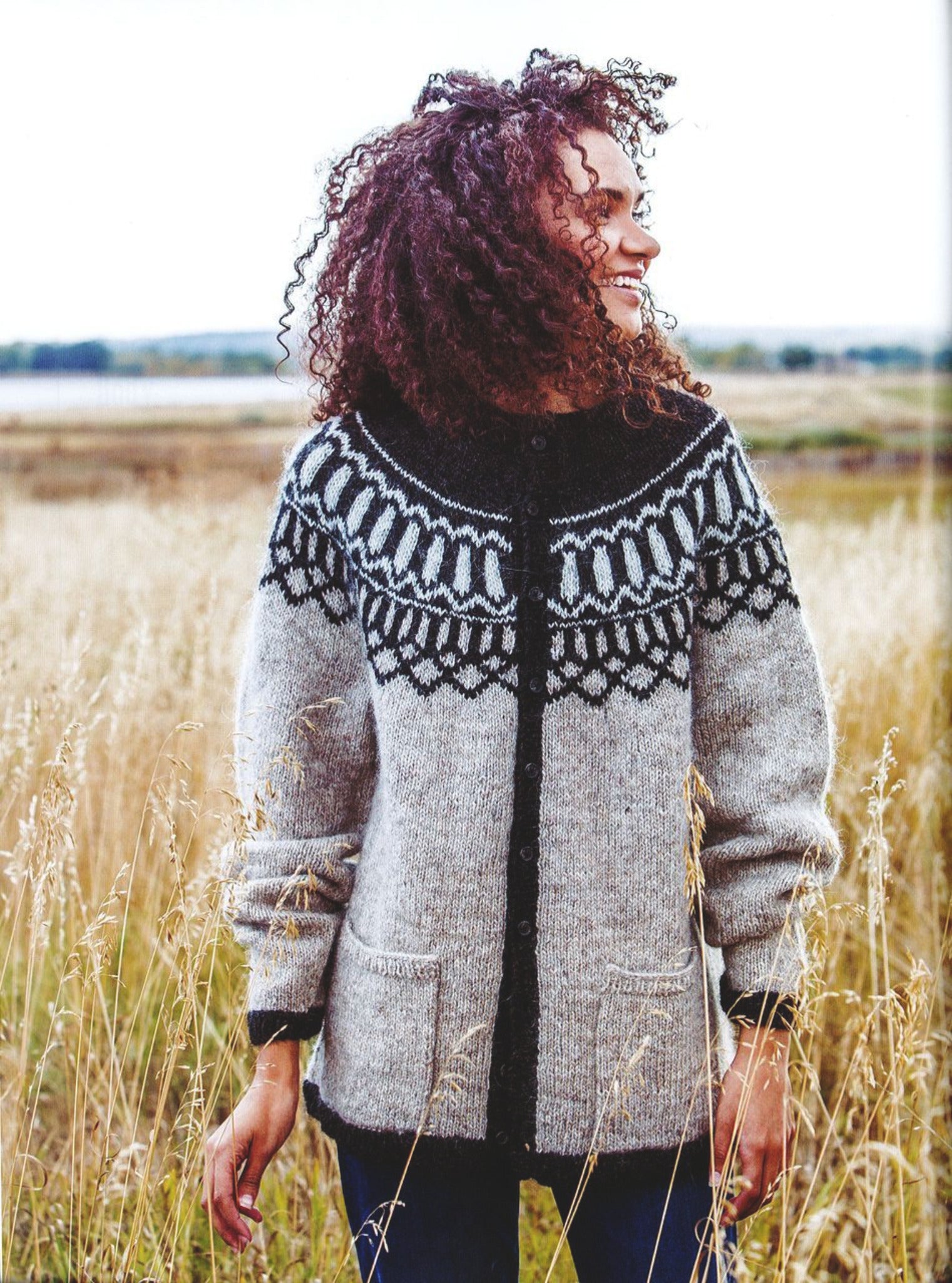 A person with curly hair stands in a field of tall grass, smiling and looking to the side. They are wearing a cozy creation from Rugged Knits - 24 Practical Projects for Everyday Living by Ingram Content, featuring an intricate black and white knit pattern across the chest and shoulders. The background is slightly blurred, emphasizing the natural scene.