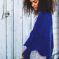 A person with curly hair is standing in front of a rustic white wooden wall, wearing an intricately patterned royal blue knitted shawl from the collection "Rugged Knits - 24 Practical Projects for Everyday Living" by Ingram Content, draped over a light gray shirt and blue jeans. They appear to be looking down thoughtfully.