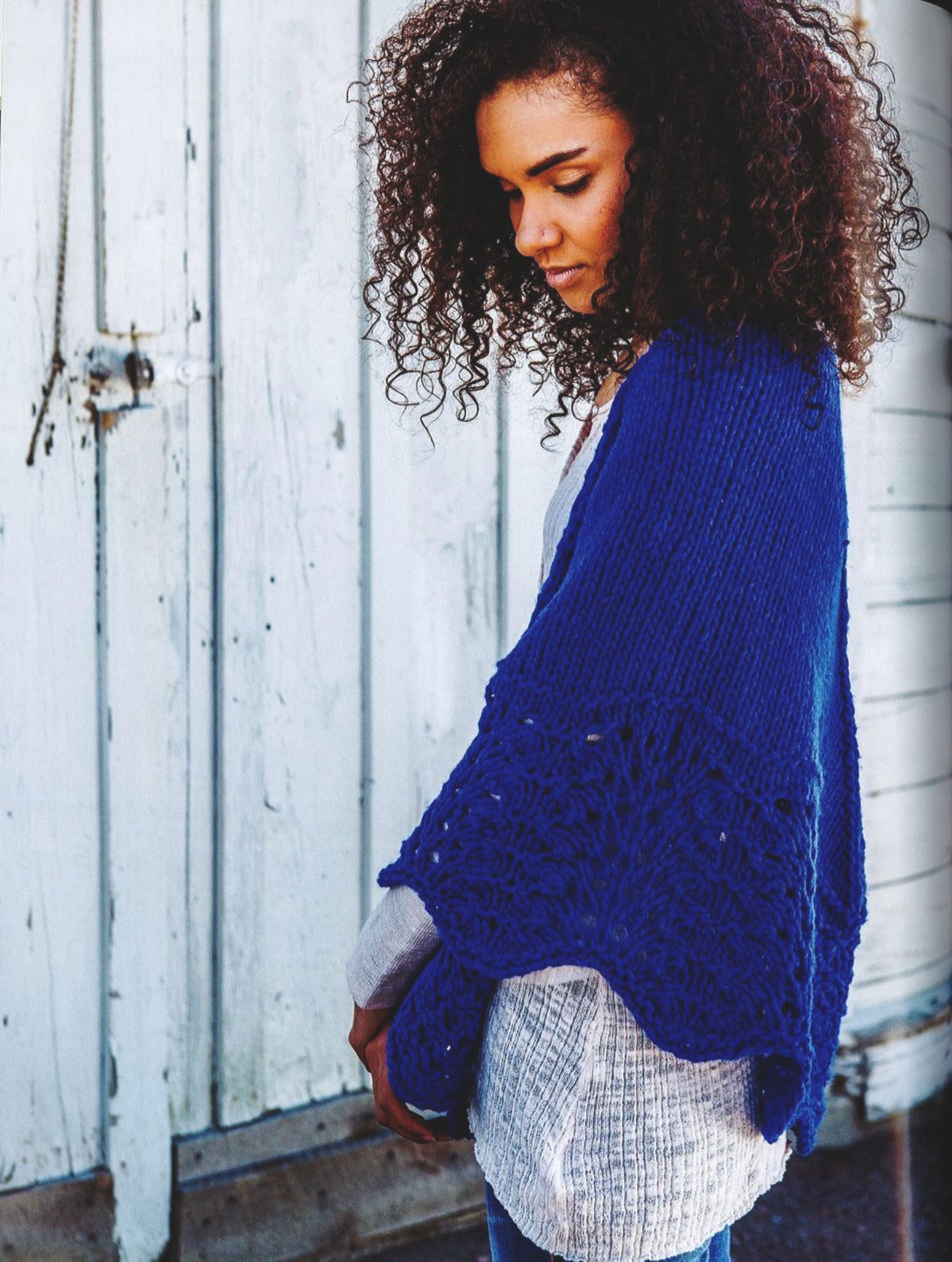 A person with curly hair is standing in front of a rustic white wooden wall, wearing an intricately patterned royal blue knitted shawl from the collection "Rugged Knits - 24 Practical Projects for Everyday Living" by Ingram Content, draped over a light gray shirt and blue jeans. They appear to be looking down thoughtfully.