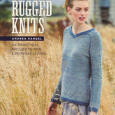 The cover of the book "Rugged Knits - 24 Practical Projects for Everyday Living" by Andrea Rangel, published by Ingram Content, showcases a woman standing in a field wearing a blue knitted sweater. The text on the cover reads "Rugged Knits" and "24 Practical Projects for Everyday Living." The background features tall grasses and blurred structures, emphasizing unique knit patterns.