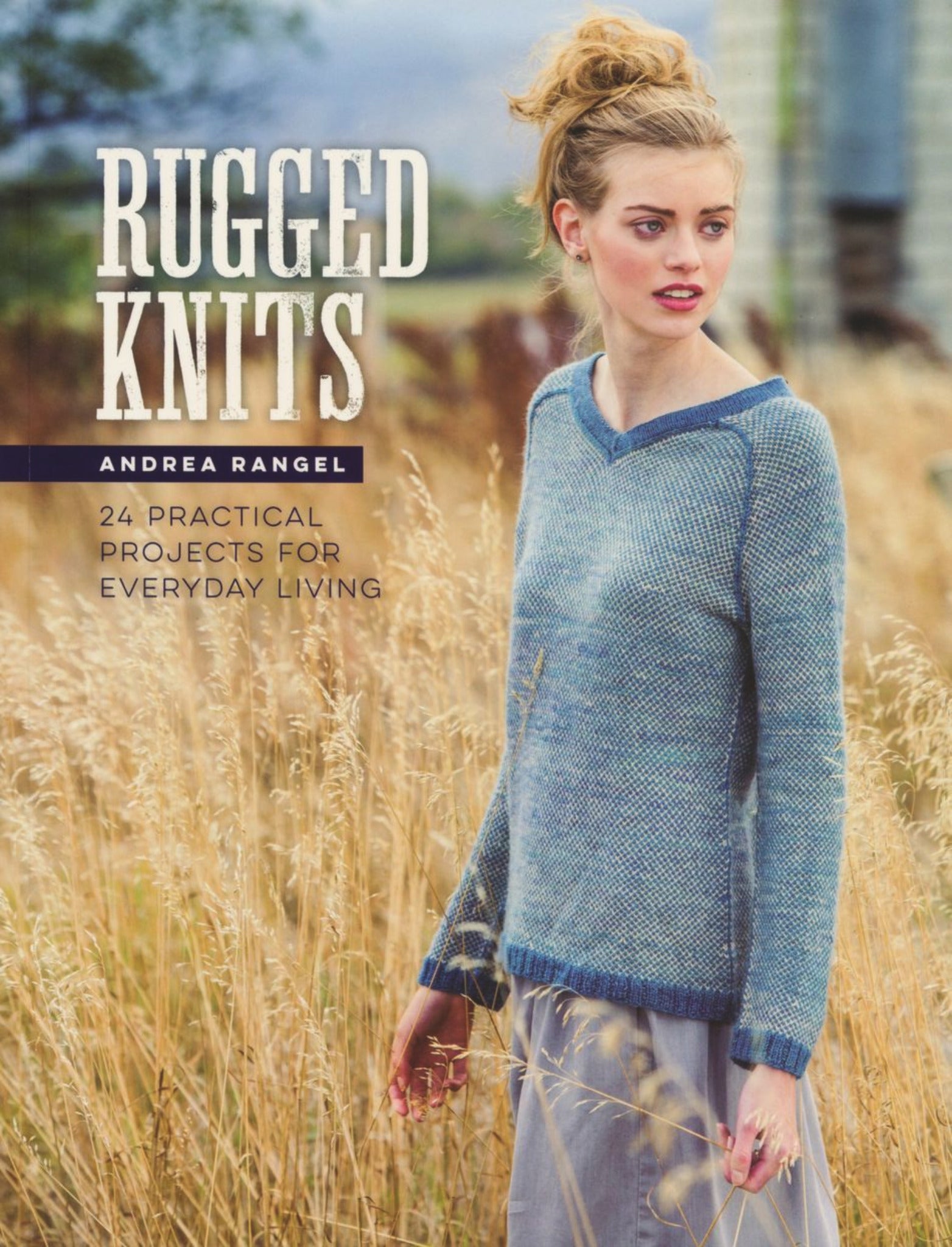 The cover of the book "Rugged Knits - 24 Practical Projects for Everyday Living" by Andrea Rangel, published by Ingram Content, showcases a woman standing in a field wearing a blue knitted sweater. The text on the cover reads "Rugged Knits" and "24 Practical Projects for Everyday Living." The background features tall grasses and blurred structures, emphasizing unique knit patterns.