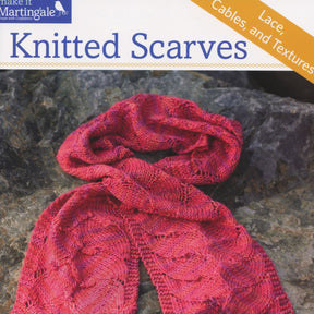 The cover of the knitting book titled "Knitted Scarves - Lace, Cables and Textures" features a pink, intricately knitted scarf laid out on a textured stone surface. Ocean-inspired designs accentuate the delicate lace patterns. The brand name, Martingale & Co, is mentioned in the top left corner.