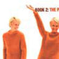 Two people are partially visible, both wearing orange sweaters. One person stands with a neutral expression while the other raises one arm. The text "The Knitting Experience Book 2: The Purl Stitch" from the learn-to-knit series by XRX is partially visible at the top right corner. The image is slightly blurry.