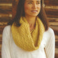 A woman with long brown hair stands in front of a wooden background, wearing a white blouse and a chunky, yellow knitted scarf with a diamond pattern. She looks off to the side with a slight smile, perfectly showcasing the chic cowls from Ingram Content's Weekend Wraps collection.