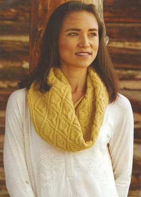 A woman with long brown hair stands in front of a wooden background, wearing a white blouse and a chunky, yellow knitted scarf with a diamond pattern. She looks off to the side with a slight smile, perfectly showcasing the chic cowls from Ingram Content's Weekend Wraps collection.