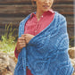 A woman wearing a red blouse stands outdoors, draped in a large, intricately patterned blue knitted Weekend Wrap from Ingram Content. She is looking down and holding the shawl with both hands. The background includes rocks and blurred greenery.