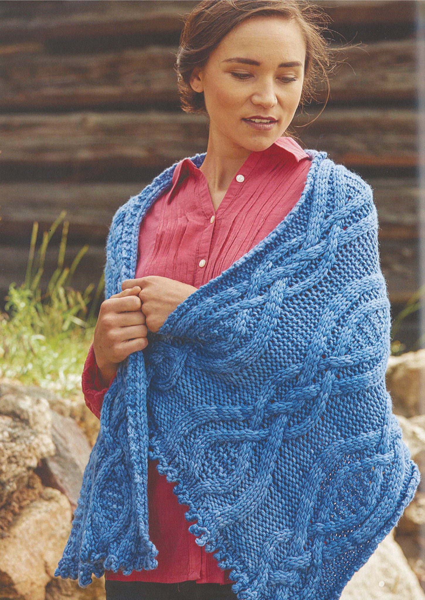 A woman wearing a red blouse stands outdoors, draped in a large, intricately patterned blue knitted Weekend Wrap from Ingram Content. She is looking down and holding the shawl with both hands. The background includes rocks and blurred greenery.