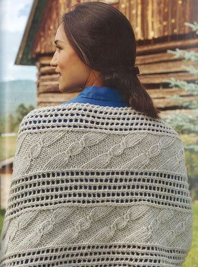 An individual with long, dark hair tied back is seen from behind. They are dressed in a blue shirt and one of Ingram Content's Weekend Wraps, showcasing a cream-colored pattern of cables and eyelets. The backdrop features a rustic wooden building and part of a green tree, evoking a rural atmosphere.