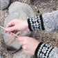 A person holding a stick over a rocky surface is wearing the "Knit Like A Latvian" grey sweater by *Ingram Content*, featuring black-and-white patterned cuffs reminiscent of traditional Latvian mitten knitting patterns. The ground beneath them is covered in small pebbles and larger rocks.