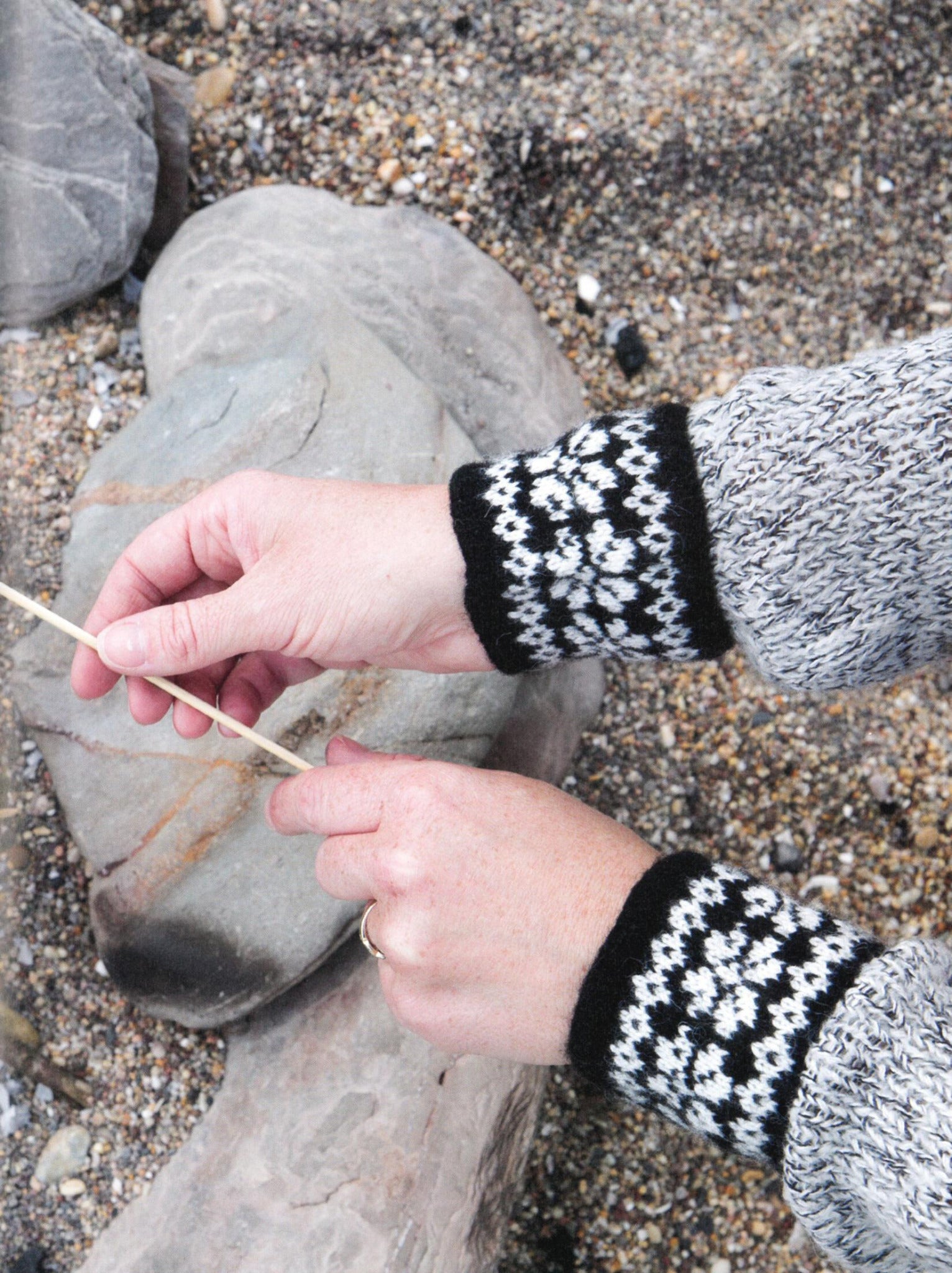 A person holding a stick over a rocky surface is wearing the "Knit Like A Latvian" grey sweater by *Ingram Content*, featuring black-and-white patterned cuffs reminiscent of traditional Latvian mitten knitting patterns. The ground beneath them is covered in small pebbles and larger rocks.