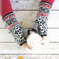 A person, adorned in a red knitted sweater and patterned black and white mittens from the "Knit Like A Latvian" collection by Ingram Content, holds a cup of hot chocolate with marshmallows on a rustic white wooden table. Two gingerbread cookies with smiling faces are displayed at the lower edge of the image.