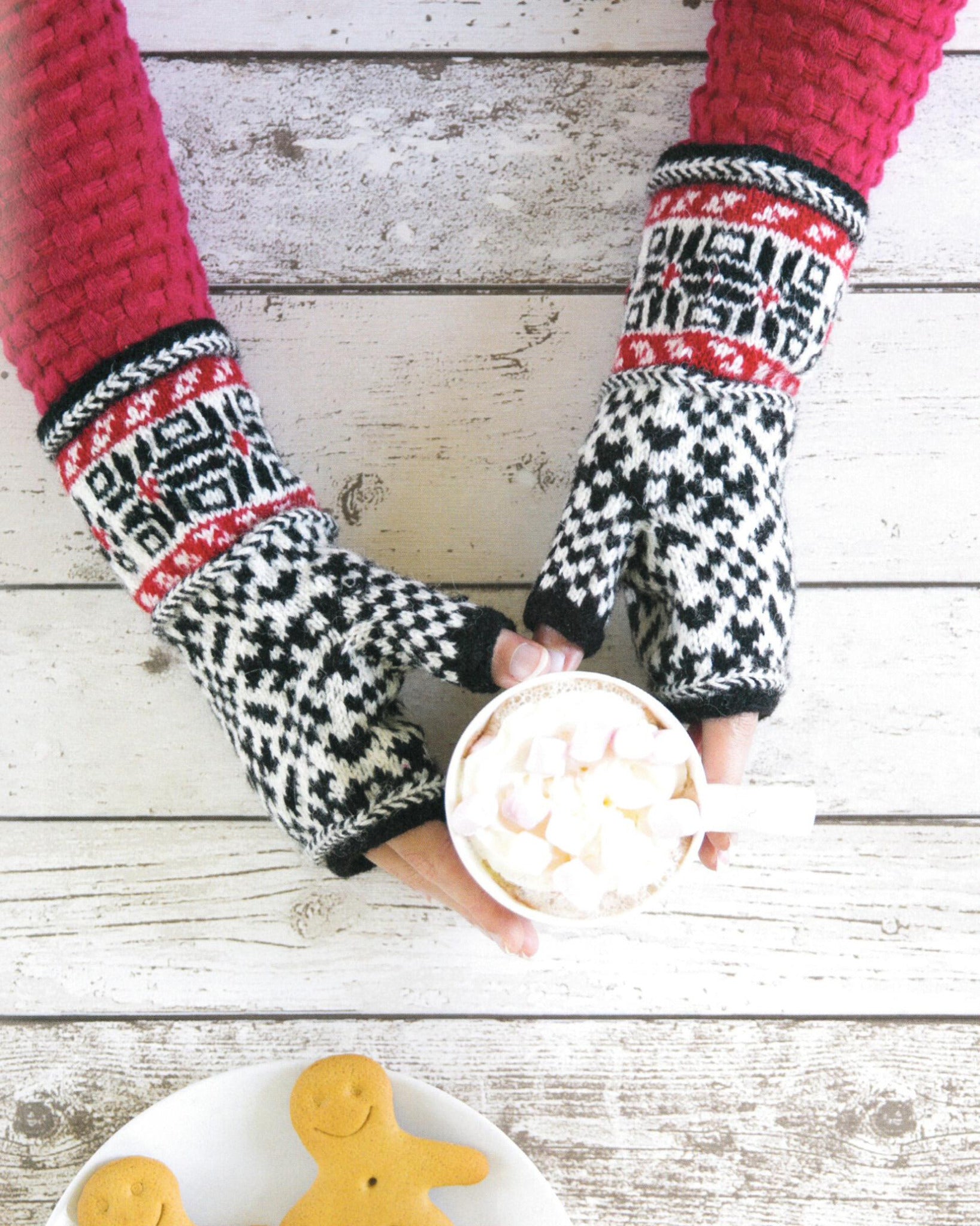 A person, adorned in a red knitted sweater and patterned black and white mittens from the "Knit Like A Latvian" collection by Ingram Content, holds a cup of hot chocolate with marshmallows on a rustic white wooden table. Two gingerbread cookies with smiling faces are displayed at the lower edge of the image.