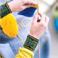 A pair of hands is seen knitting with yellow yarn, wearing a yellow long-sleeve shirt and wristbands featuring colorful traditional designs. The project showcases blue and yellow sections on a light blue textured background, utilizing Latvian mitten knitting patterns from "Knit Like A Latvian" by Ingram Content to create intricate fingerless gloves.