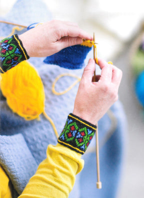 A pair of hands is seen knitting with yellow yarn, wearing a yellow long-sleeve shirt and wristbands featuring colorful traditional designs. The project showcases blue and yellow sections on a light blue textured background, utilizing Latvian mitten knitting patterns from "Knit Like A Latvian" by Ingram Content to create intricate fingerless gloves.