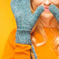 A woman with long, wavy hair is standing against an orange background, wearing a bright orange sweater and teal Knit Mitts by Ingram Content. She is playfully holding the mittens up to her nose and smiling.