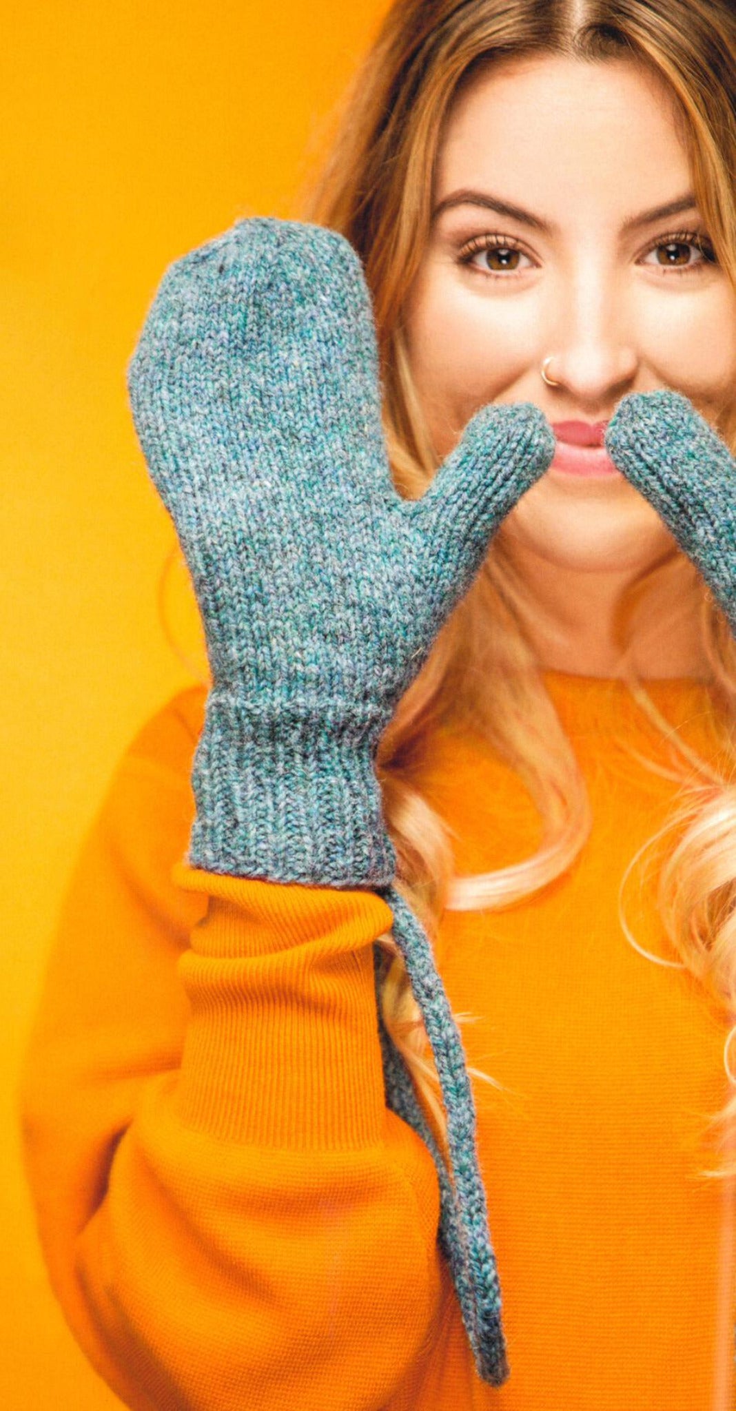 A woman with long, wavy hair is standing against an orange background, wearing a bright orange sweater and teal Knit Mitts by Ingram Content. She is playfully holding the mittens up to her nose and smiling.