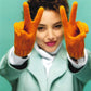 A cheerful person with curly hair, wearing an aqua coat and bright orange mittens from "Knit Mitts - Your Hand-Y Guide to Knitting Mittens & Gloves" by Ingram Content, stands against a green background. They hold up both hands in front of them, each making a peace sign.