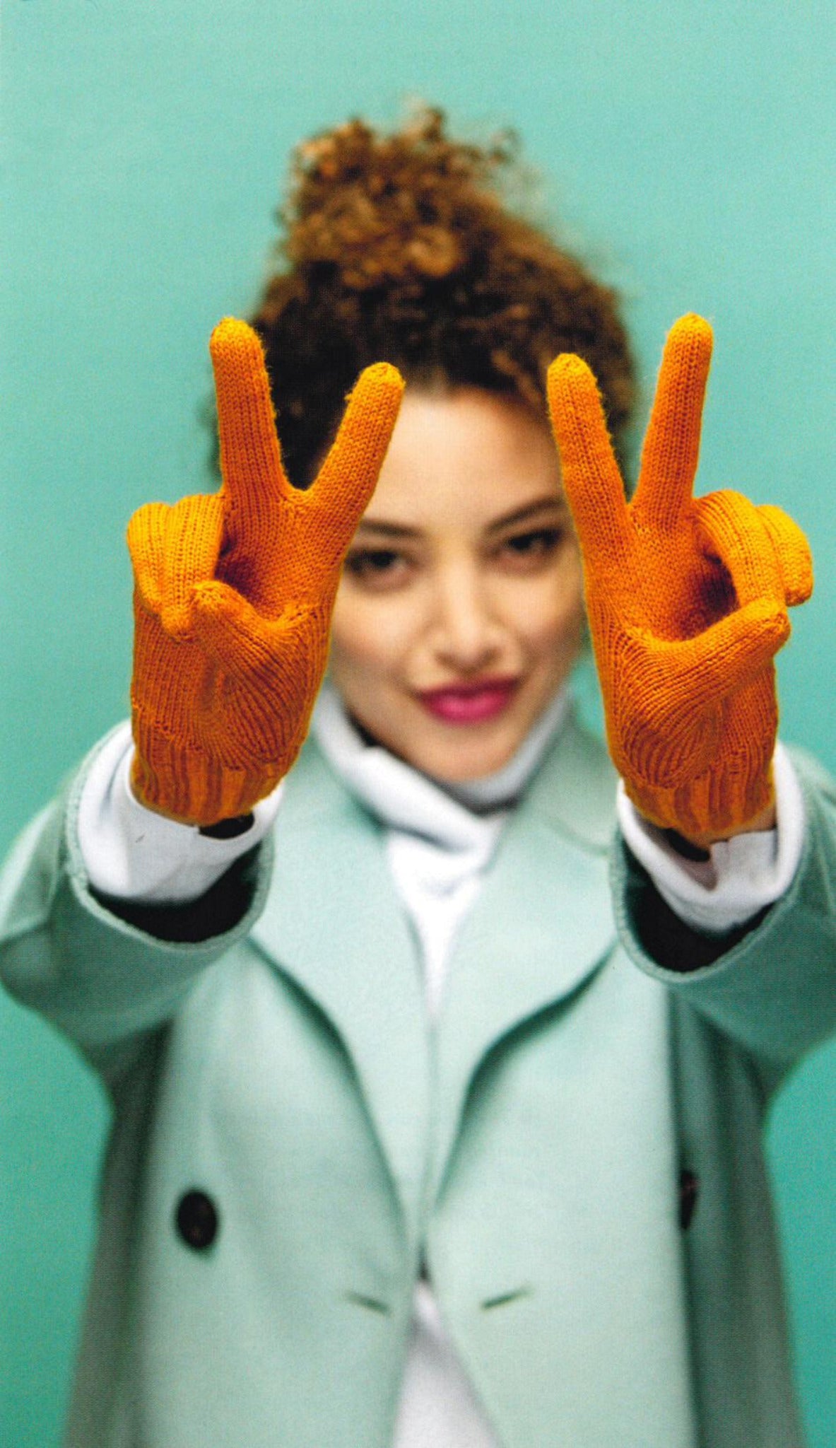 A cheerful person with curly hair, wearing an aqua coat and bright orange mittens from "Knit Mitts - Your Hand-Y Guide to Knitting Mittens & Gloves" by Ingram Content, stands against a green background. They hold up both hands in front of them, each making a peace sign.