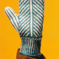 A person wearing a brown coat is holding up their hand, showcasing a mitten knitted using patterns from "Knit Mitts - Your Hand-Y Guide to Knitting Mittens & Gloves" by Ingram Content. The expert creation features alternating light blue and white stripes and is set against a bright yellow background.