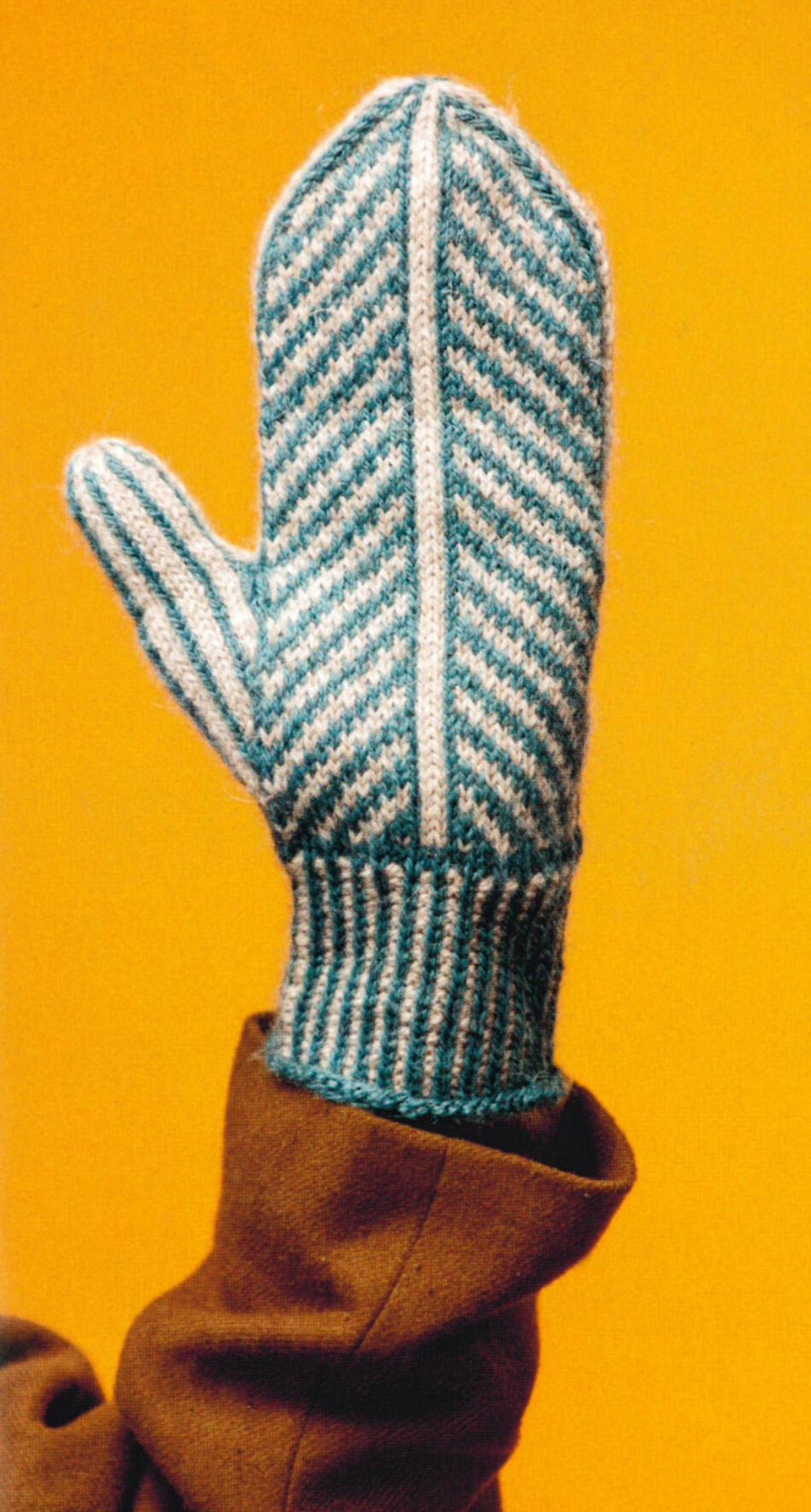 A person wearing a brown coat is holding up their hand, showcasing a mitten knitted using patterns from "Knit Mitts - Your Hand-Y Guide to Knitting Mittens & Gloves" by Ingram Content. The expert creation features alternating light blue and white stripes and is set against a bright yellow background.