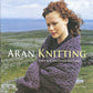 The cover of the book "Aran Knitting - New and Expanded Edition" by Alice Starmore, published by Ingram Content, showcases a woman with curly red hair wearing a cable-knit sweater. She stands outdoors against a backdrop of rocky terrain and cloudy skies, embodying the rich tradition of charted patterns in Aran knitting.