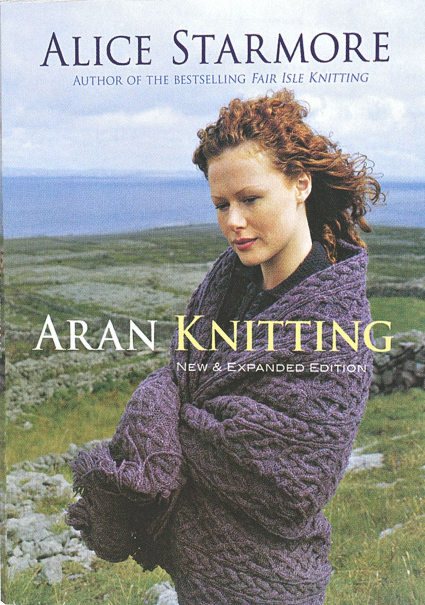 The cover of the book "Aran Knitting - New and Expanded Edition" by Alice Starmore, published by Ingram Content, showcases a woman with curly red hair wearing a cable-knit sweater. She stands outdoors against a backdrop of rocky terrain and cloudy skies, embodying the rich tradition of charted patterns in Aran knitting.