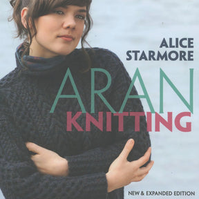 A woman with dark hair, in a braid wrapped over her head, is wearing a thick, dark-blue, cable-knit sweater. Her arms are crossed. Text on the image reads, "Aran Knitting - New and Expanded Edition" by Ingram Content, showcasing charted patterns in this essential knitting book.
