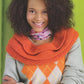 A person with curly hair smiles while wearing a light gray sweater with an orange argyle pattern and R & M West Coast Enterprises' "Cowl Girls" large orange chunky cowl draped over their shoulders. They also have a pink patterned scarf around their neck and a bracelet adorned with Swarovski crystals on their wrist, standing against a green background.