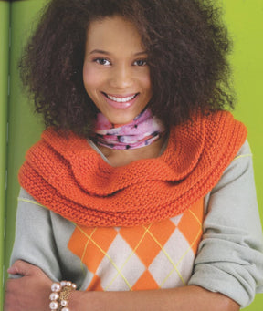 A person with curly hair smiles while wearing a light gray sweater with an orange argyle pattern and R & M West Coast Enterprises' "Cowl Girls" large orange chunky cowl draped over their shoulders. They also have a pink patterned scarf around their neck and a bracelet adorned with Swarovski crystals on their wrist, standing against a green background.