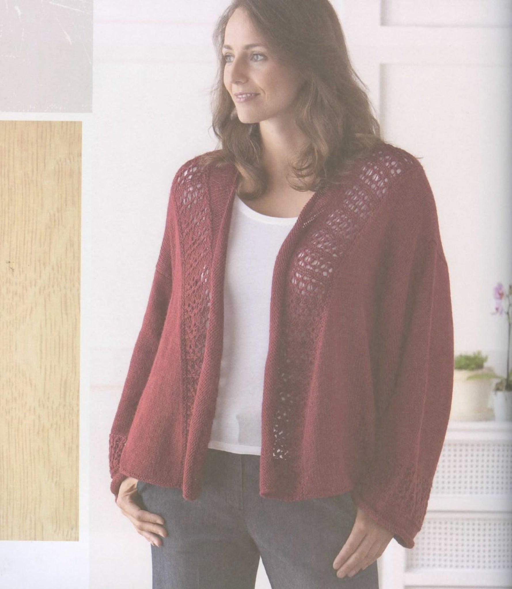 A woman with long brown hair stands indoors wearing a deep red Knitting Green cardigan by Ingram Content, made from organic yarn, over a white top and dark jeans. She is looking slightly to her right and smiling. The background features neutral tones and minimalist decor, including a wooden panel and a white planter.