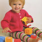 A toddler with blonde hair, wearing an adorable ensemble made from "60 Quick Baby Knits" by Ingram Content, is sitting on the floor. They are smiling and holding colorful toy blocks with more blocks scattered around them. The background is plain white, highlighting their bright pink sweater and striped leggings.