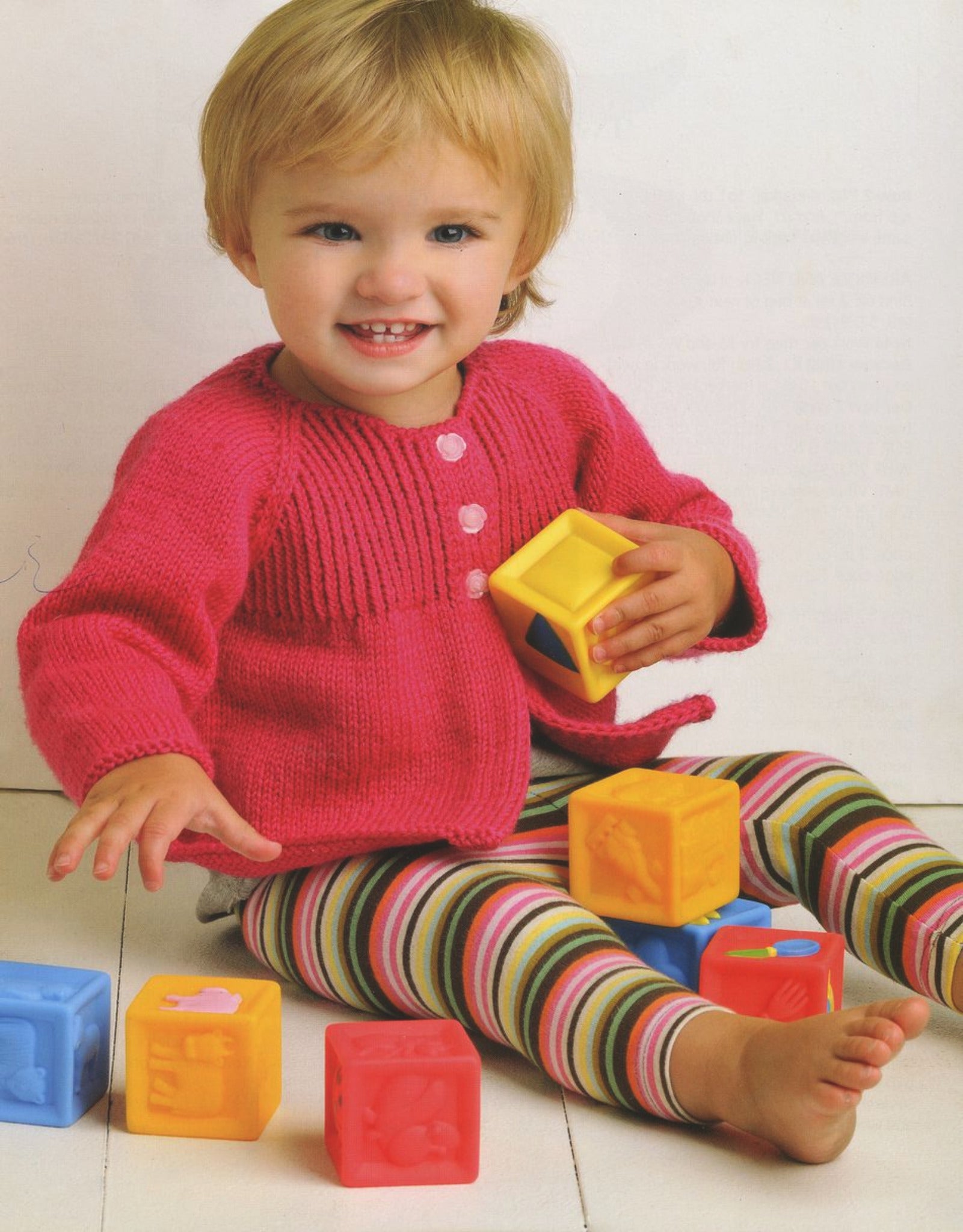 A toddler with blonde hair, wearing an adorable ensemble made from "60 Quick Baby Knits" by Ingram Content, is sitting on the floor. They are smiling and holding colorful toy blocks with more blocks scattered around them. The background is plain white, highlighting their bright pink sweater and striped leggings.