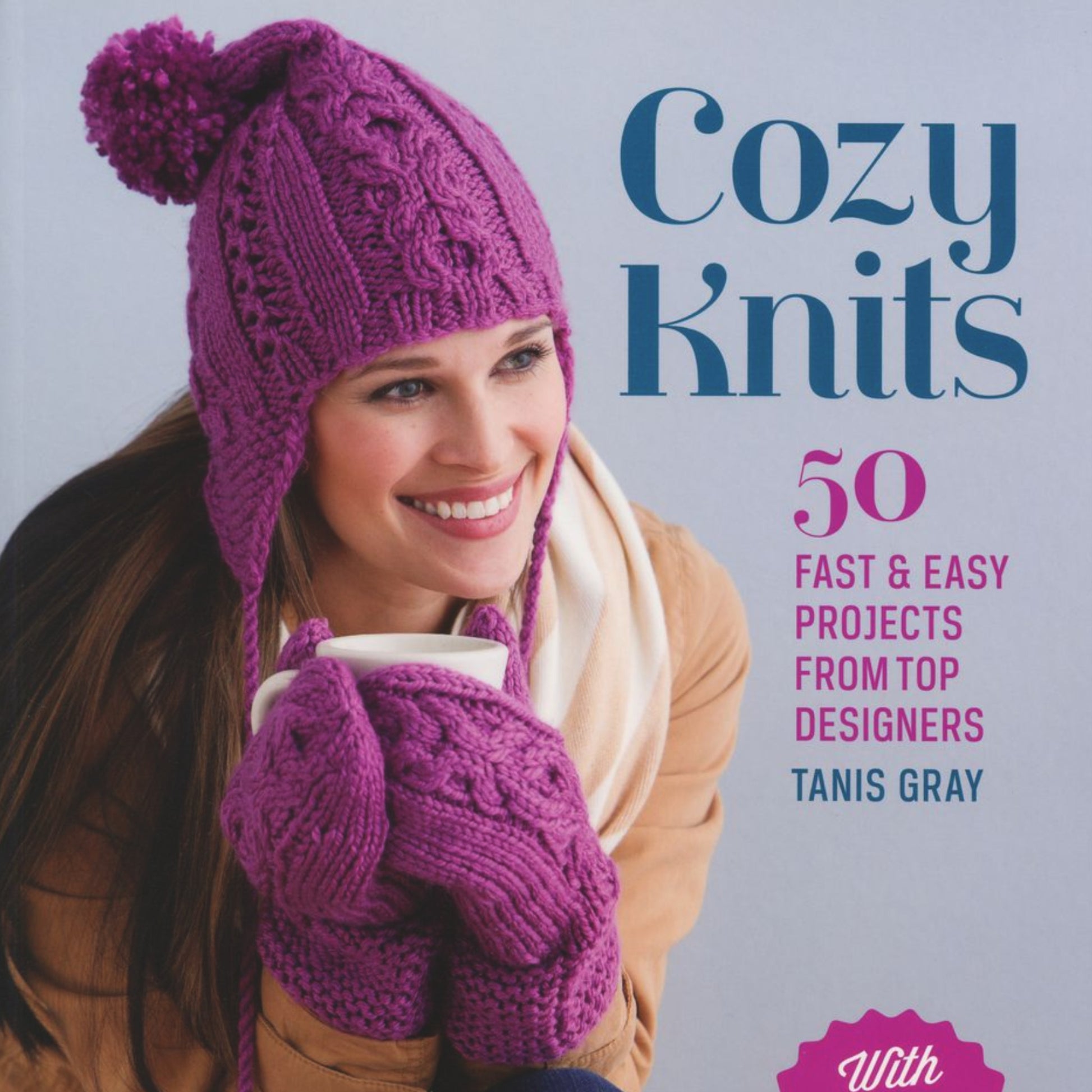 A woman bundled in a coat, wearing a purple knit hat and mittens, smiles as she holds a white mug. The text reads, "Cozy Knits" by Ingram Content. She embodies a warm, cozy atmosphere perfect for knitting projects using worsted and bulky yarns.