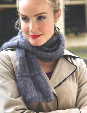 A woman with light hair tied up is wearing a beige trench coat and a gray scarf, which complements her red lipstick and slight smile. She accessorizes with a large earring that appears to be black, white, and gold, exuding elegance reminiscent of the sophisticated style highlighted in "Metropolitan Knits - Chic Designs for Urban Style" by Ingram Content. The background is slightly blurred.