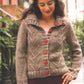 A woman stands outdoors on a city street, showcasing an urban-style sweater from "Metropolitan Knits - Chic Designs for Urban Style" by Ingram Content. The knitted taupe cardigan features a herringbone pattern and red buttons. She has long, dark hair and is looking down with a gentle smile. The background reveals a railing, trees, and residential buildings.