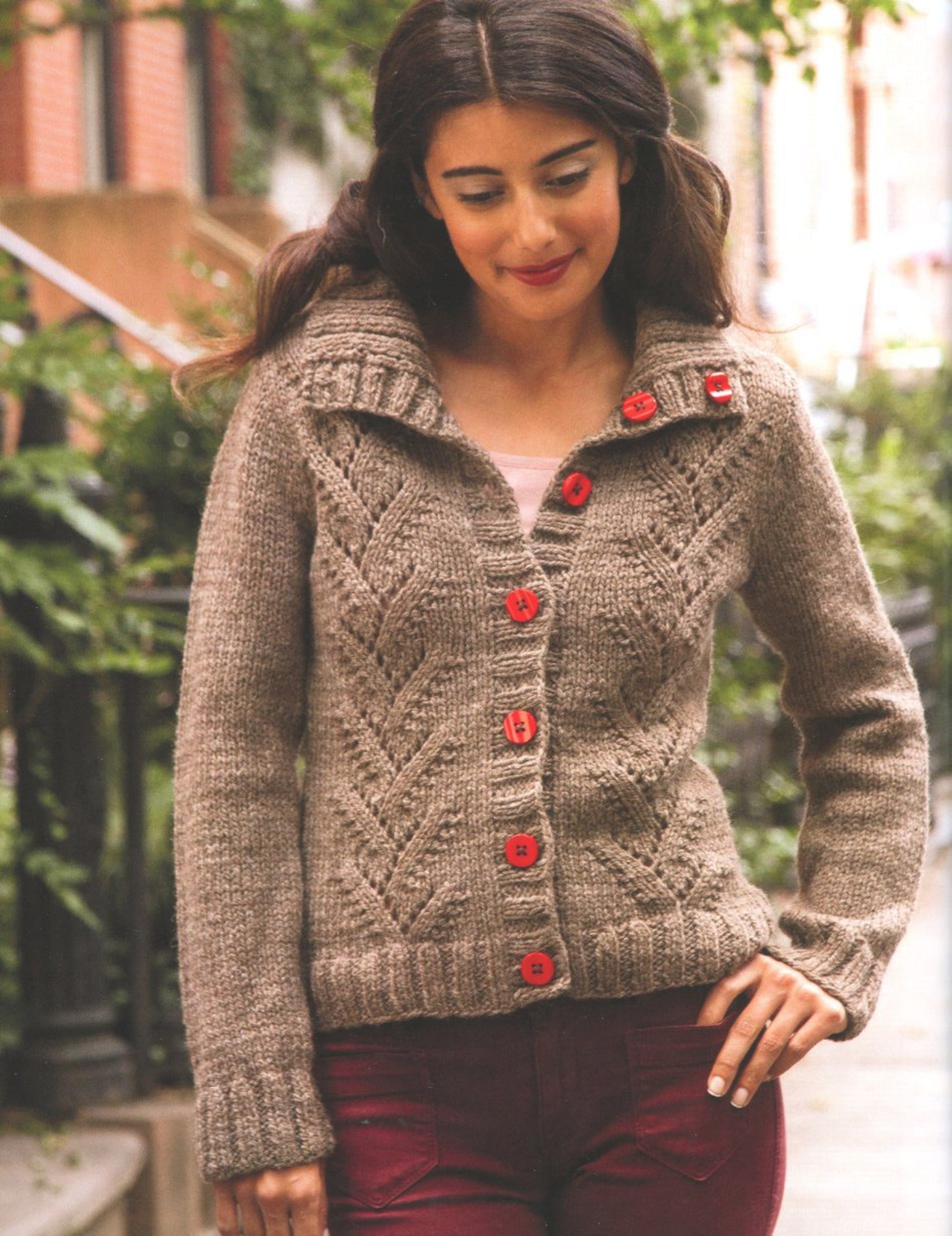 A woman stands outdoors on a city street, showcasing an urban-style sweater from "Metropolitan Knits - Chic Designs for Urban Style" by Ingram Content. The knitted taupe cardigan features a herringbone pattern and red buttons. She has long, dark hair and is looking down with a gentle smile. The background reveals a railing, trees, and residential buildings.